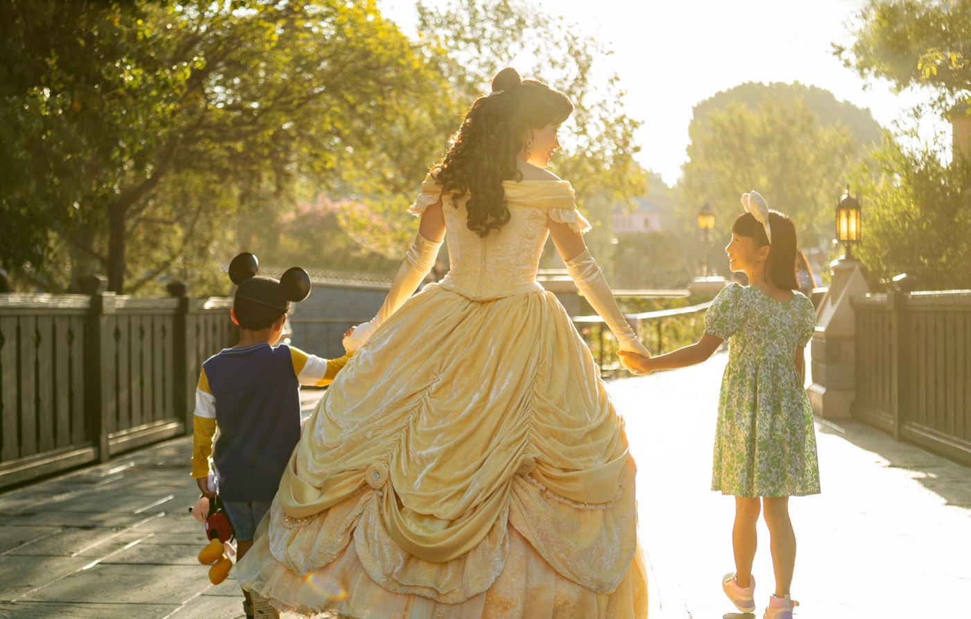 Belle walking with two children through a sunlit Disney park.